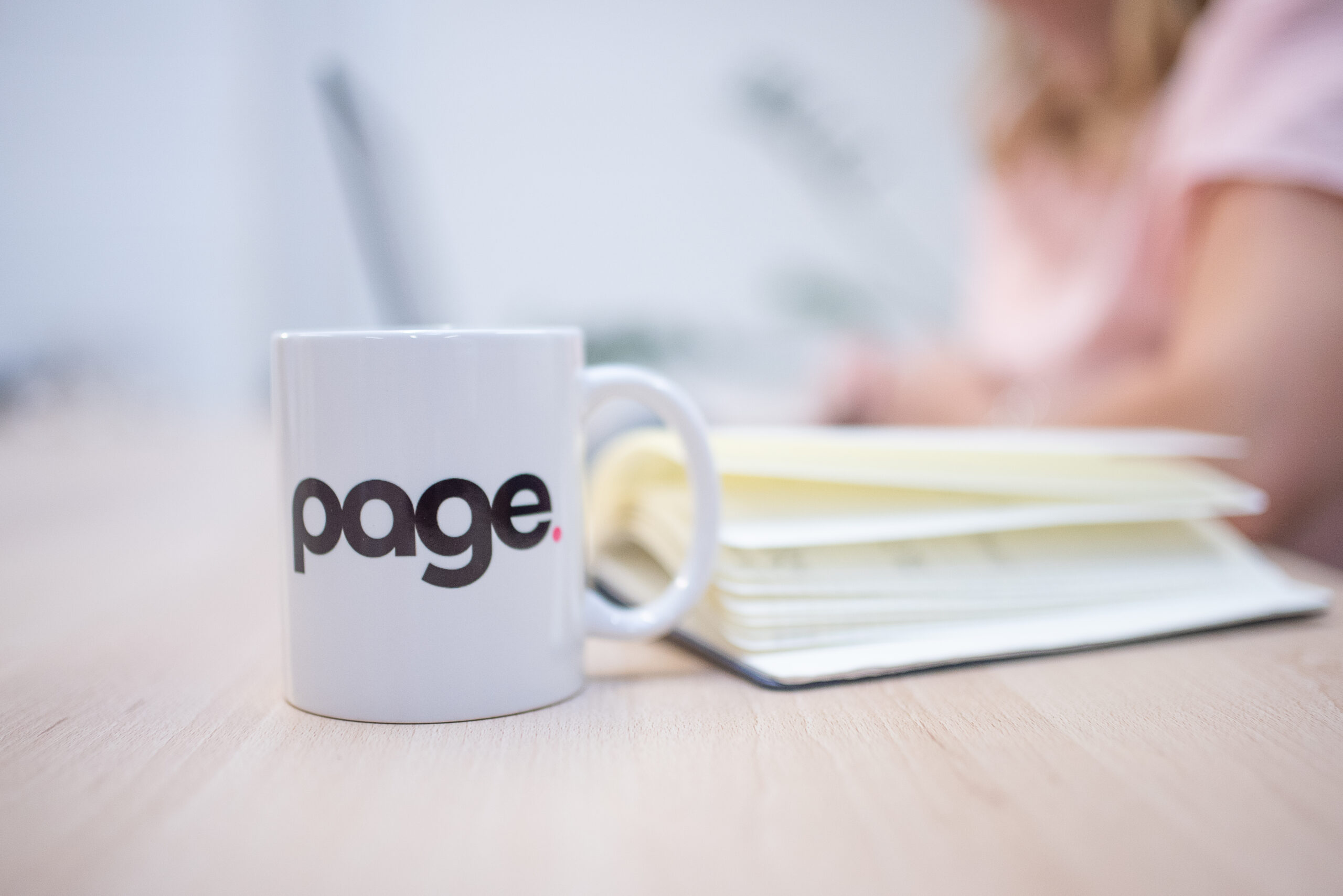 Page mug in the foreground with notepad and person blurred in the background