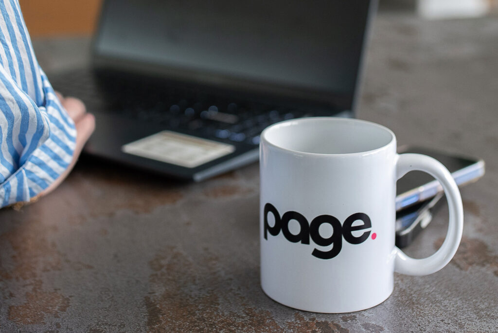 WHITE MUG WITH PAGE WRITTEN ON IT WITH LAPTOP IN BACKGROUND