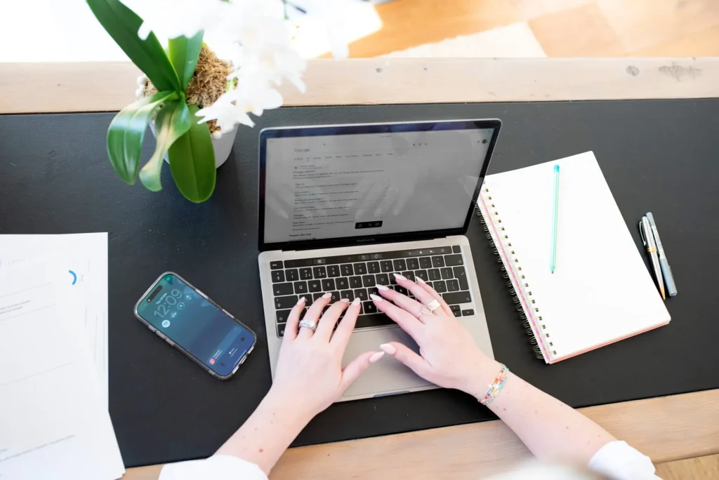 Google on a laptop with a phone and notepad either side of the laptop. Hands are typing on the keyboard